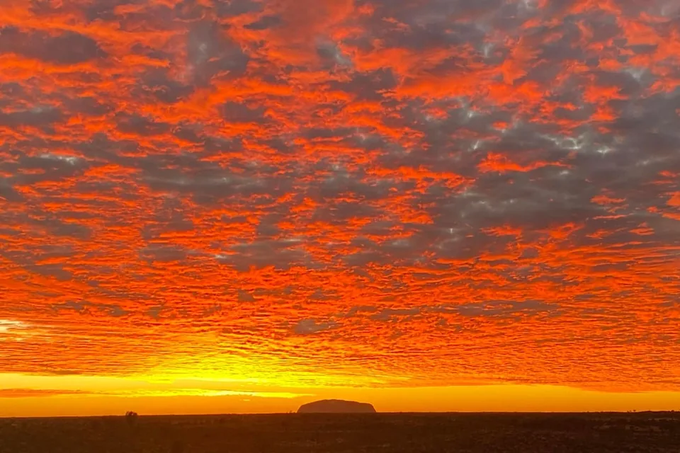 A fiery orange sunset sky over the desert with Uluru a small silhouette on the horizon